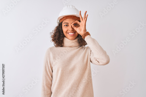 Young brazilian architect woman wearing security helmet over isolated white background doing ok gesture with hand smiling, eye looking through fingers with happy face.