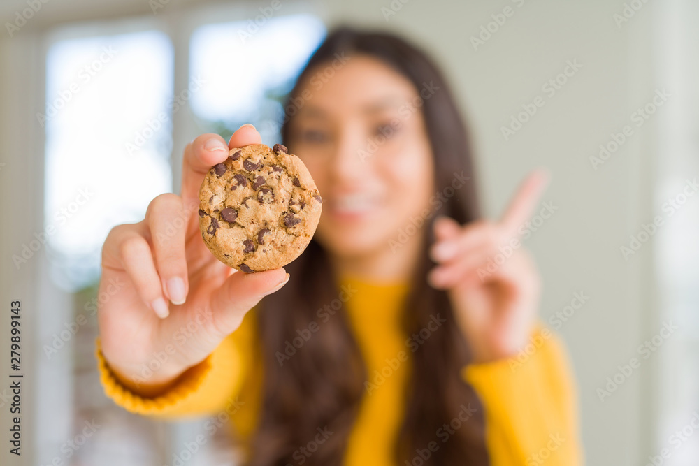 Young woman eating chocolate chips cookies at home surprised with an idea or question pointing finger with happy face, number one Young woman eating chocolate chips cookies at home surprised with an idea or question pointing finger with happy face, number one