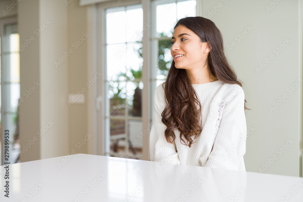 Young beautiful woman at home on white table looking away to side with smile on face, natural expression. Laughing confident.