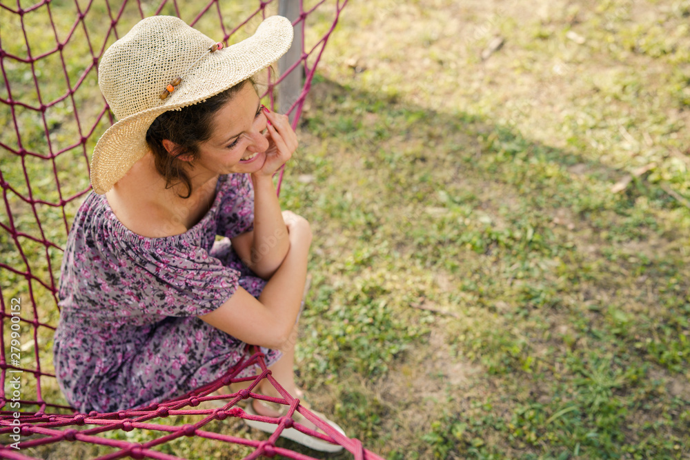 Obraz premium Young woman holding her summer hat while sitting in net hammock in her backyard in sunny day smiling happy