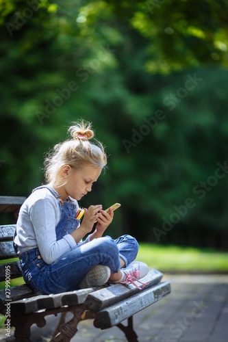 Happy Little beautiful school girl in elementary school age sitting on a park bench and looking at mobile phone outdoors on summer day.