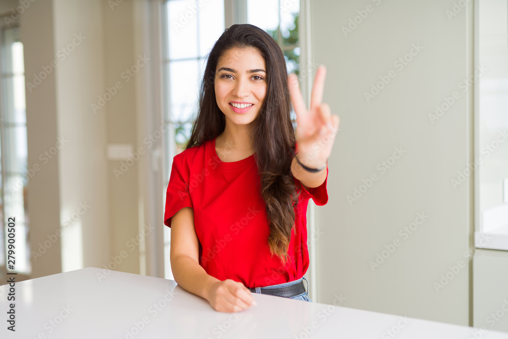 Young beautiful woman at home on white table smiling with happy face winking at the camera doing victory sign. Number two. Young beautiful woman at home on white table smiling with happy face winking at the camera doing victory sign. Number two.