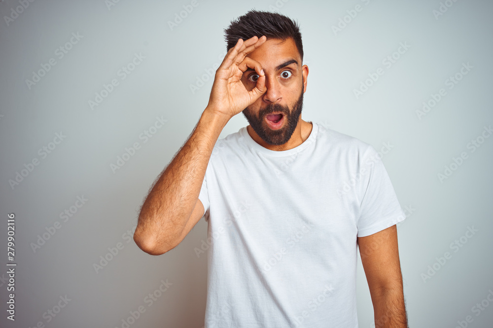 Young indian man wearing t-shirt standing over isolated white background doing ok gesture shocked with surprised face, eye looking through fingers. Unbelieving expression. Young indian man wearing t-shirt standing over isolated white background doing ok gesture shocked with surprised face, eye looking through fingers. Unbelieving expression.