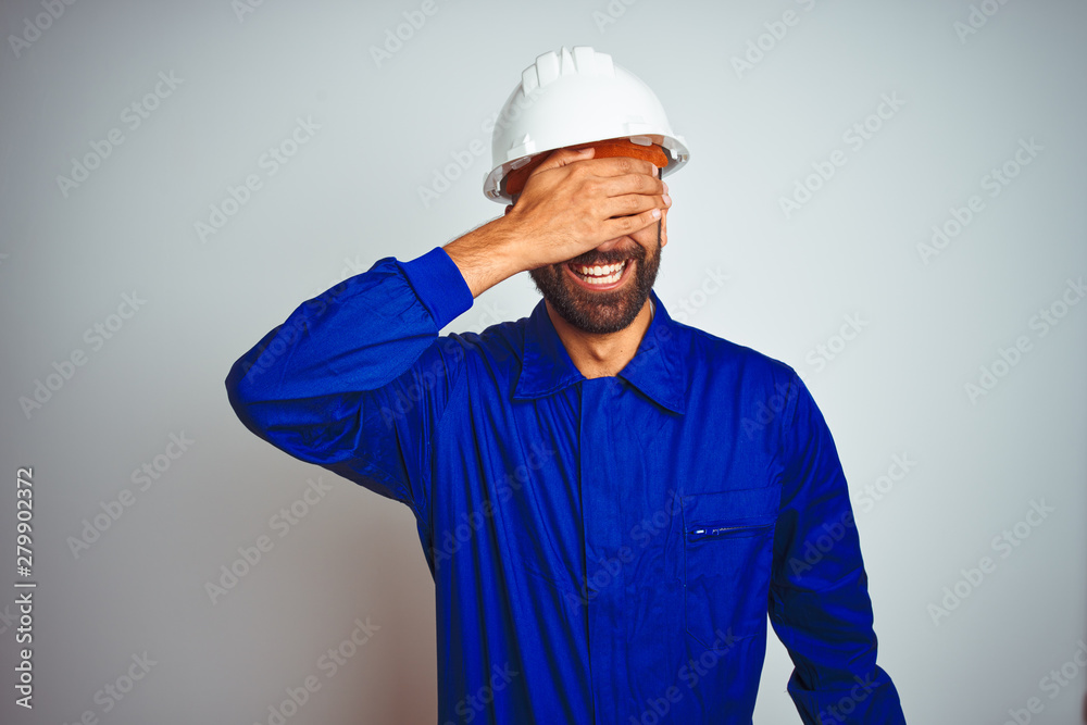 Handsome indian worker man wearing uniform and helmet over isolated white background smiling and laughing with hand on face covering eyes for surprise. Blind concept.