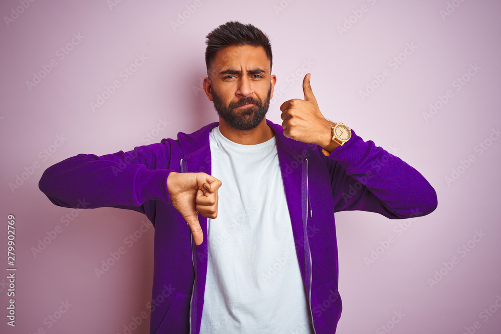 Young indian man wearing purple sweatshirt standing over isolated pink background Doing thumbs up and down, disagreement and agreement expression. Crazy conflict Young indian man wearing purple sweatshirt standing over isolated pink background Doing thumbs up and down, disagreement and agreement expression. Crazy conflict