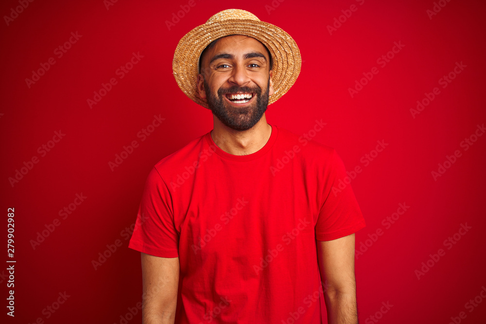 Young handsome indian man wearing t-shirt and hat over isolated red background with a happy and cool smile on face. Lucky person.