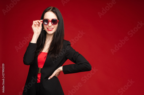 Portrait of young beautiful woman thoughtfully looking down. Brunette model in elegant costume posing at camera . Pretty lady in red sunglasses leaning one hand on other and holding it near face. © Вячеслав Косько