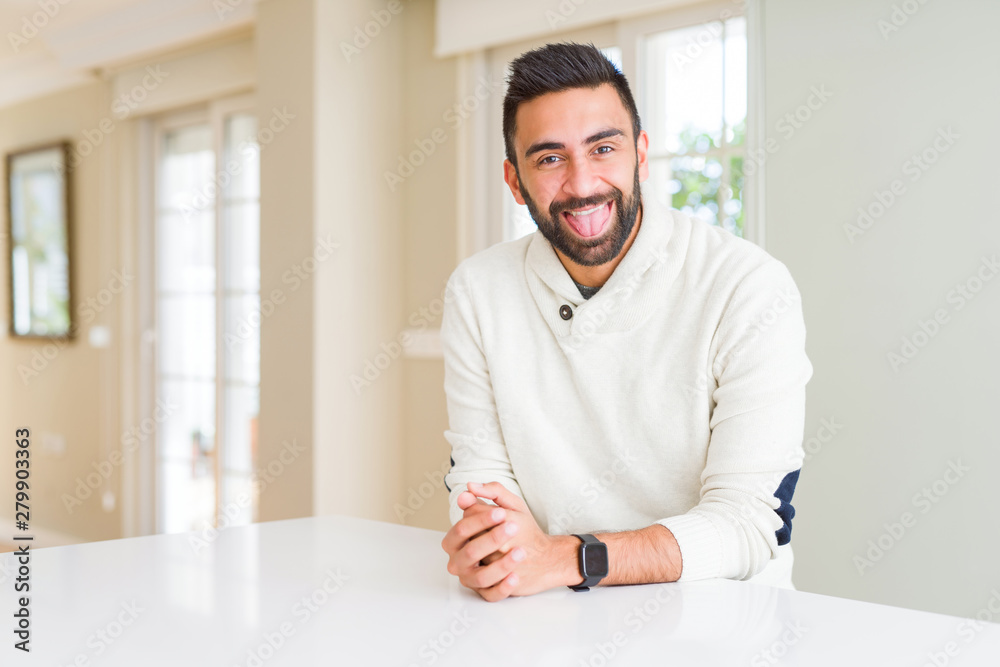 Fototapeta premium Handsome hispanic man wearing casual white sweater at home sticking tongue out happy with funny expression. Emotion concept.