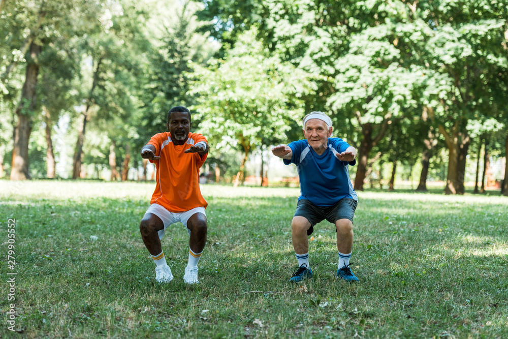 happy senior and multicultural med doing sit ups on grass near trees happy senior and multicultural med doing sit ups on grass near trees