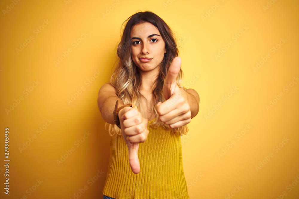 Young beautiful woman wearing t-shirt over yellow isolated background Doing thumbs up and down, disagreement and agreement expression. Crazy conflict Young beautiful woman wearing t-shirt over yellow isolated background Doing thumbs up and down, disagreement and agreement expression. Crazy conflict