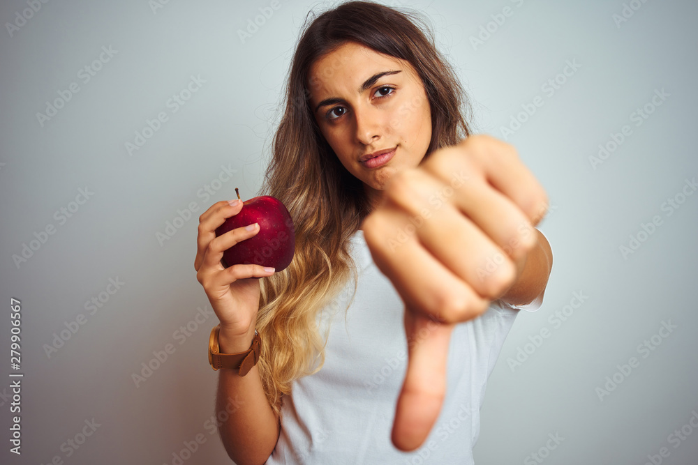 Young beautiful woman eating red apple over grey isolated background with angry face, negative sign showing dislike with thumbs down, rejection concept Young beautiful woman eating red apple over grey isolated background with angry face, negative sign showing dislike with thumbs down, rejection concept