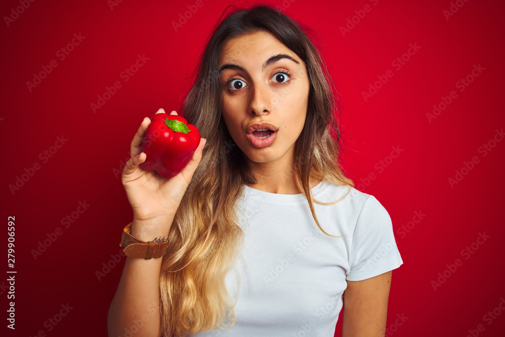 Young beautiful woman holding pepper over red isolated background scared in shock with a surprise face, afraid and excited with fear expression Young beautiful woman holding pepper over red isolated background scared in shock with a surprise face, afraid and excited with fear expression
