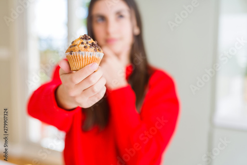 Beautiful young woman eating chocolate chips muffin serious face thinking about question  very confused idea