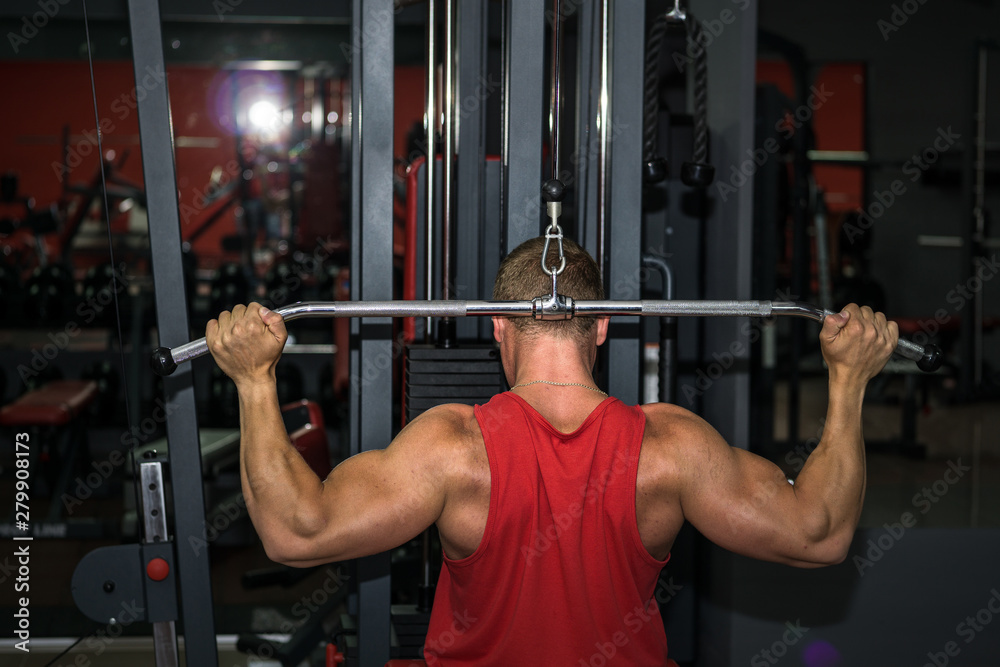Muscular young man doing exercices on exercise machine in the gym. View from the back. Sport, lifestyle, training and health care concept.