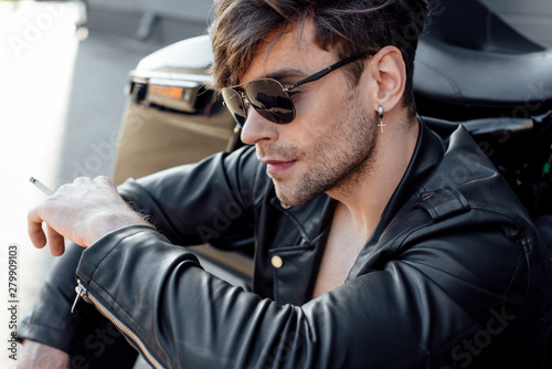 young motorcyclist in leather jacket smoking while sitting near motorcycle