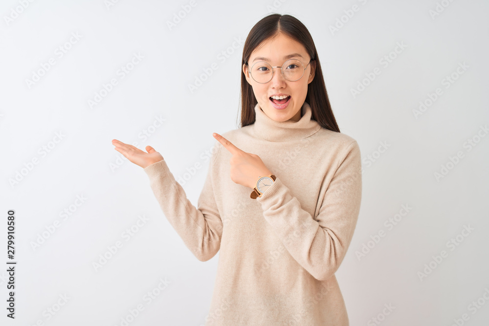Young chinese woman wearing turtleneck sweater and glasses over isolated white background amazed and smiling to the camera while presenting with hand and pointing with finger. Young chinese woman wearing turtleneck sweater and glasses over isolated white background amazed and smiling to the camera while presenting with hand and pointing with finger.