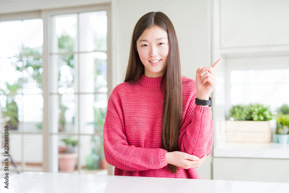 Beautiful Asian woman wearing pink sweater on white table with a big smile on face, pointing with hand and finger to the side looking at the camera.