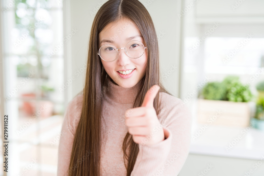 Beautiful Asian woman wearing glasses doing happy thumbs up gesture with hand. Approving expression looking at the camera showing success.