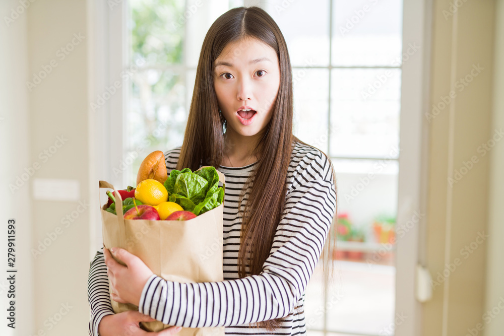Beautiful Asian woman holding paper bag of fresh groceries scared in shock with a surprise face, afraid and excited with fear expression Beautiful Asian woman holding paper bag of fresh groceries scared in shock with a surprise face, afraid and excited with fear expression