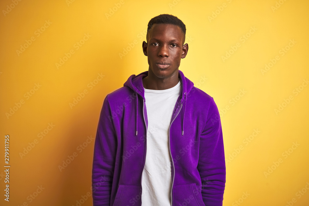 African american man wearing purple sweatshirt standing over isolated yellow background with serious expression on face. Simple and natural looking at the camera. African american man wearing purple sweatshirt standing over isolated yellow background with serious expression on face. Simple and natural looking at the camera.