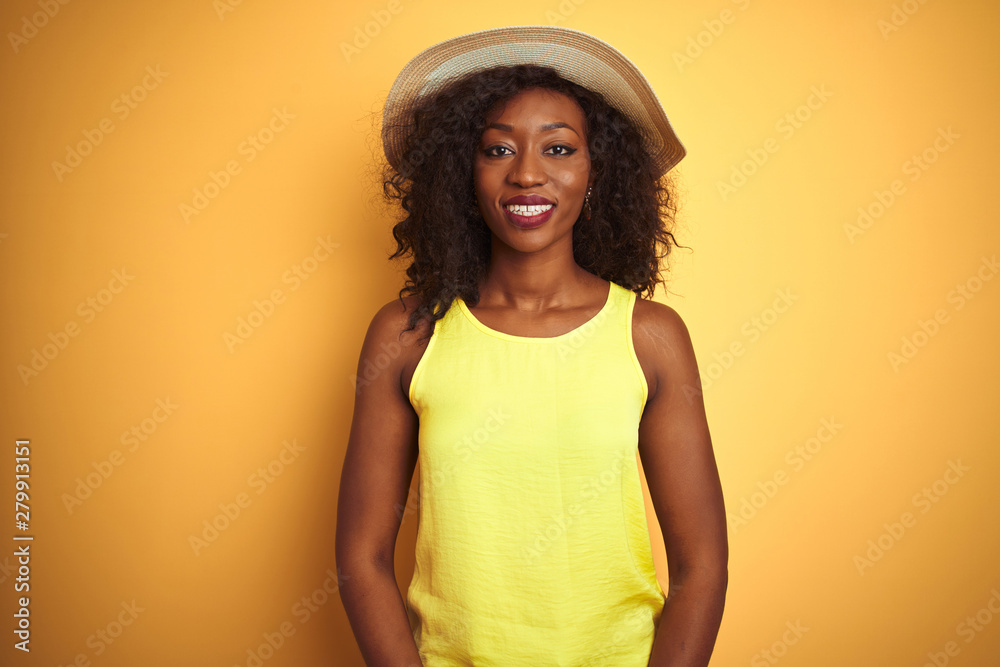 Young african american woman wearing t-shirt and hat over isolated yellow background with a happy and cool smile on face. Lucky person. Young african american woman wearing t-shirt and hat over isolated yellow background with a happy and cool smile on face. Lucky person.