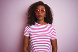Young african american woman wearing t-shirt and sunglasses over isolated pink background with serious expression on face. Simple and natural looking at the camera.