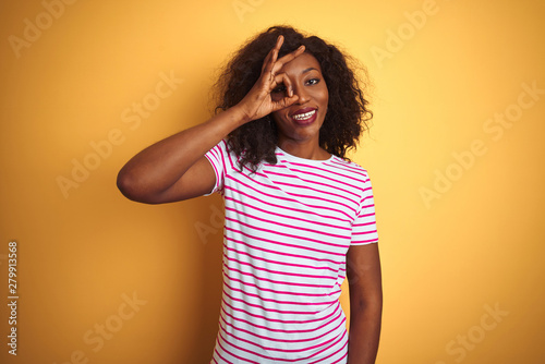 Young african american woman wearing striped t-shirt over isolated yellow background doing ok gesture with hand smiling, eye looking through fingers with happy face.