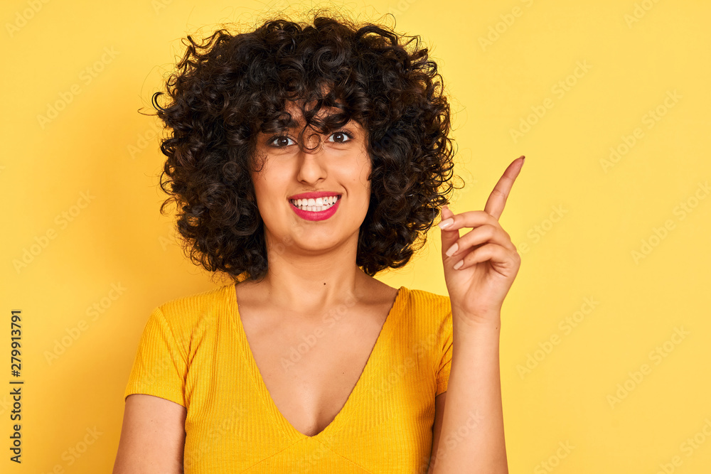 Young arab woman with curly hair wearing t-shirt standing over isolated yellow background very happy pointing with hand and finger to the side Young arab woman with curly hair wearing t-shirt standing over isolated yellow background very happy pointing with hand and finger to the side