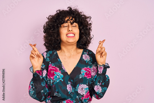 Young arab woman with curly hair wearing floral dress over isolated pink background gesturing finger crossed smiling with hope and eyes closed. Luck and superstitious concept.