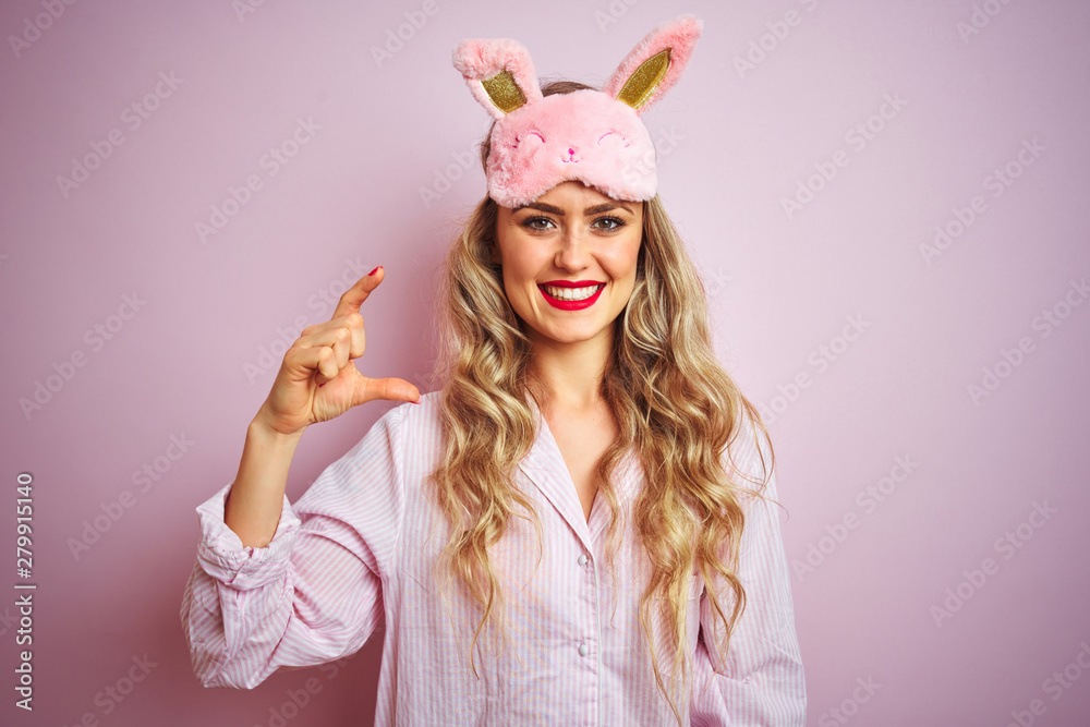 Young beautiful woman wearing pajama and sleep mask over pink isolated background smiling and confident gesturing with hand doing small size sign with fingers looking and the camera. Measure concept.