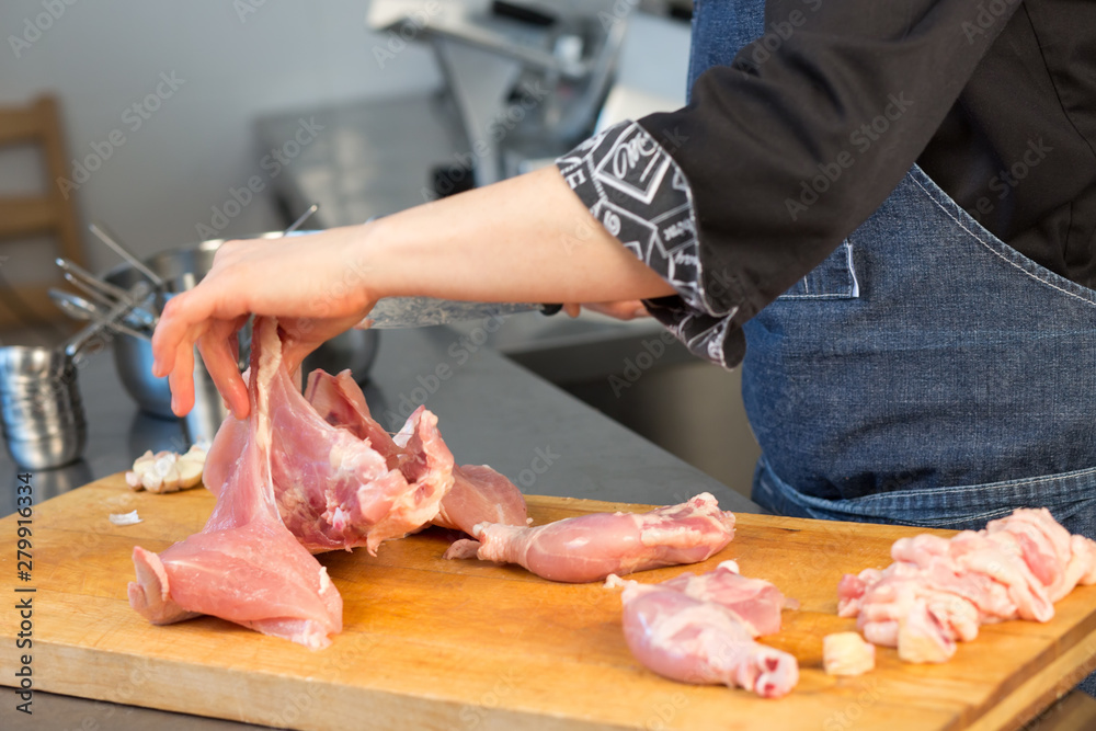 A person cuts raw chicken. Cook's hand with a knife close-up on the background of the kitchen. the woman  professional chef holds raw chicken. The background is blurred