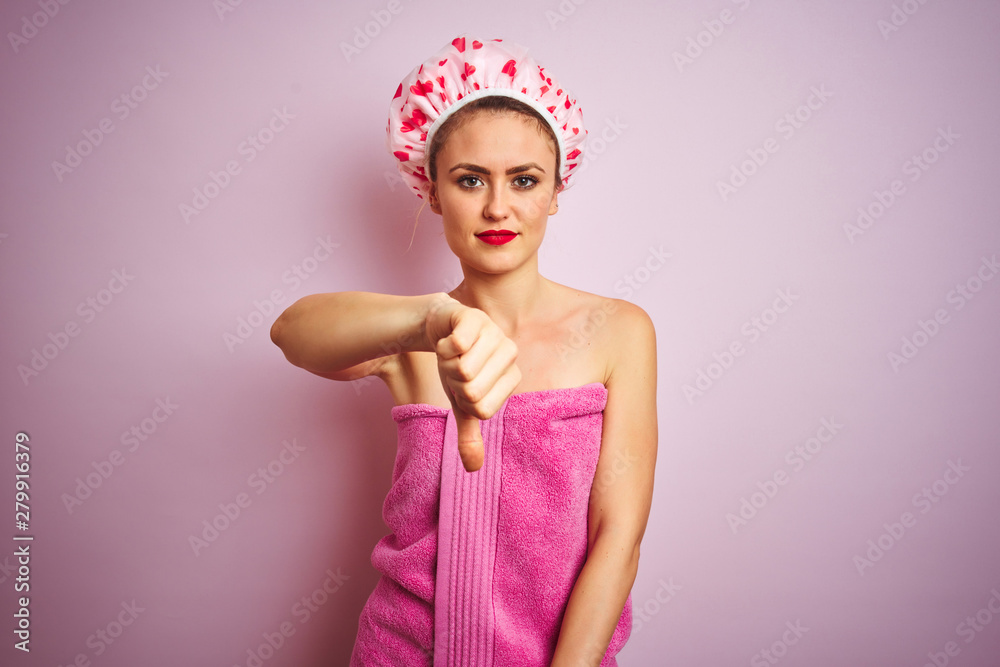 Young beautiful woman wearing towel and bath hat after shower over pink isolated background looking unhappy and angry showing rejection and negative with thumbs down gesture. Bad expression. Young beautiful woman wearing towel and bath hat after shower over pink isolated background looking unhappy and angry showing rejection and negative with thumbs down gesture. Bad expression.