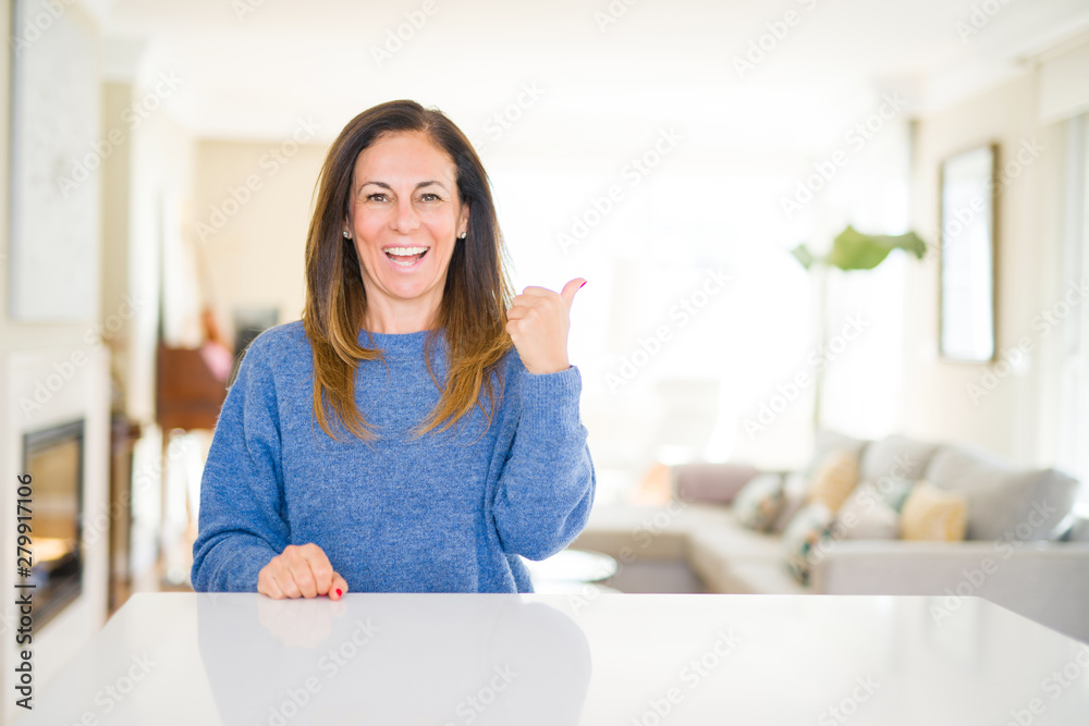 Beautiful middle age woman at home smiling with happy face looking and pointing to the side with thumb up. Beautiful middle age woman at home smiling with happy face looking and pointing to the side with thumb up.