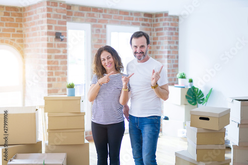 Middle age senior couple moving to a new home with boxes around smiling and looking at the camera pointing with two hands and fingers to the side.