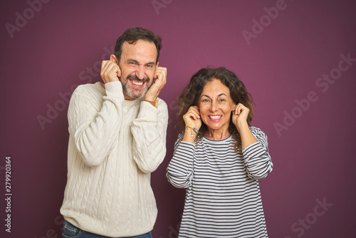 Beautiful middle age couple wearing winter sweater over isolated purple background covering ears with fingers with annoyed expression for the noise of loud music. Deaf concept.