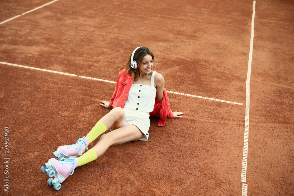Happy stylish young woman with vintage roller skates and headphones sitting on tennis court