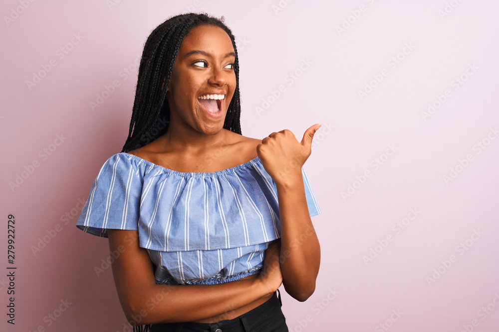 African american woman wearing striped t-shirt standing over isolated pink background smiling with happy face looking and pointing to the side with thumb up. African american woman wearing striped t-shirt standing over isolated pink background smiling with happy face looking and pointing to the side with thumb up.