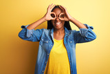 Young african american woman wearing denim shirt standing over isolated yellow background doing ok gesture like binoculars sticking tongue out, eyes looking through fingers. Crazy expression.