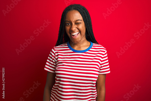 Young african american woman wearing striped t-shirt standing over isolated red background winking looking at the camera with sexy expression, cheerful and happy face.