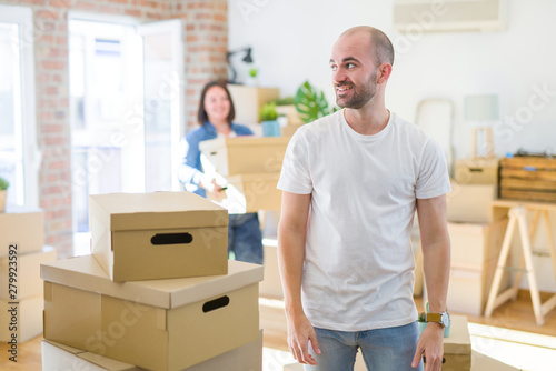Young couple arround cardboard boxes moving to a new house, bald man standing at home looking away to side with smile on face, natural expression. Laughing confident.