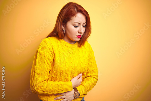 Beautiful redhead woman wearing winter sweater standing over isolated yellow background with hand on stomach because indigestion, painful illness feeling unwell. Ache concept.