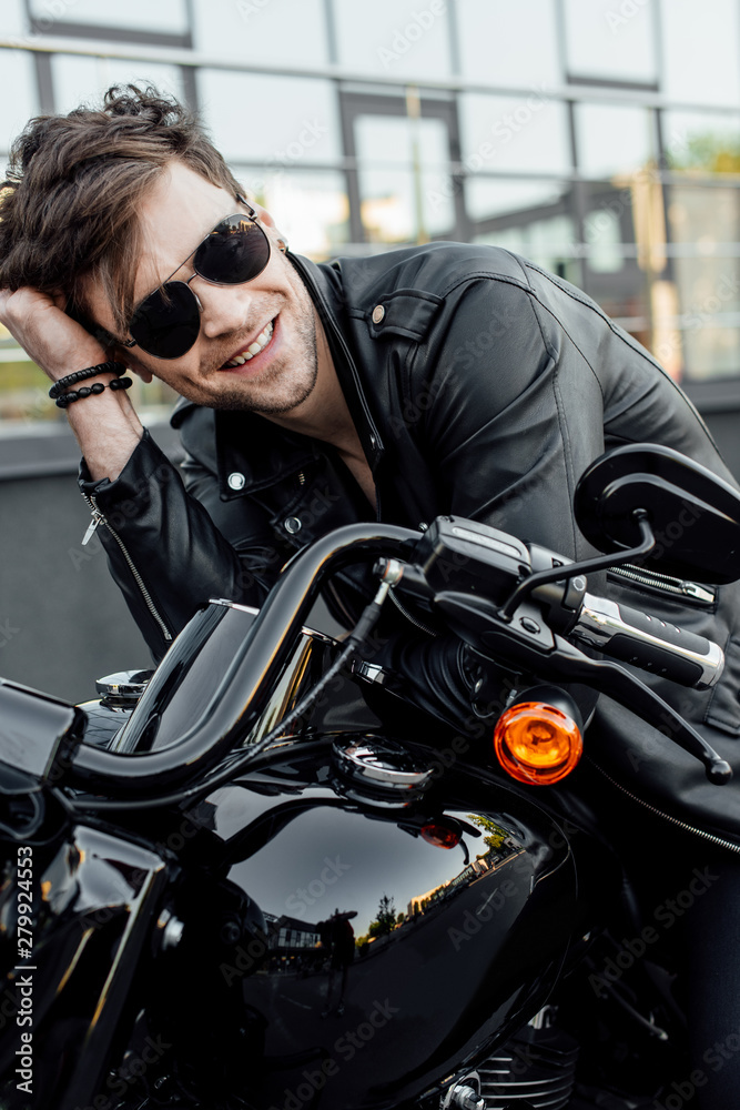 handsome young motorcyclist in sunglasses holding head with hand, smiling and looking away while sitting on motorcycle