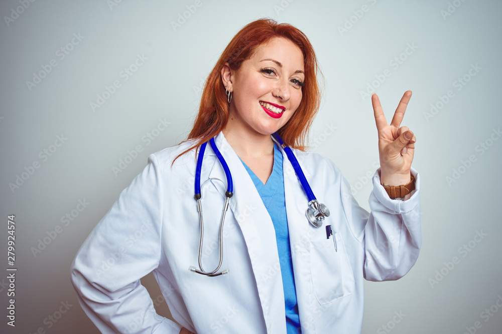Young redhead doctor woman using stethoscope over white isolated background smiling looking to the camera showing fingers doing victory sign. Number two. Young redhead doctor woman using stethoscope over white isolated background smiling looking to the camera showing fingers doing victory sign. Number two.