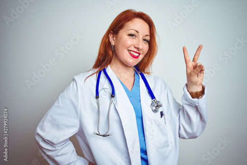 Young redhead doctor woman using stethoscope over white isolated background smiling looking to the camera showing fingers doing victory sign. Number two.