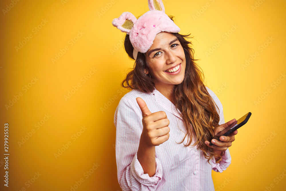 Young woman wearing pajama and mask using smartphone over yellow isolated background happy with big smile doing ok sign, thumb up with fingers, excellent sign Young woman wearing pajama and mask using smartphone over yellow isolated background happy with big smile doing ok sign, thumb up with fingers, excellent sign