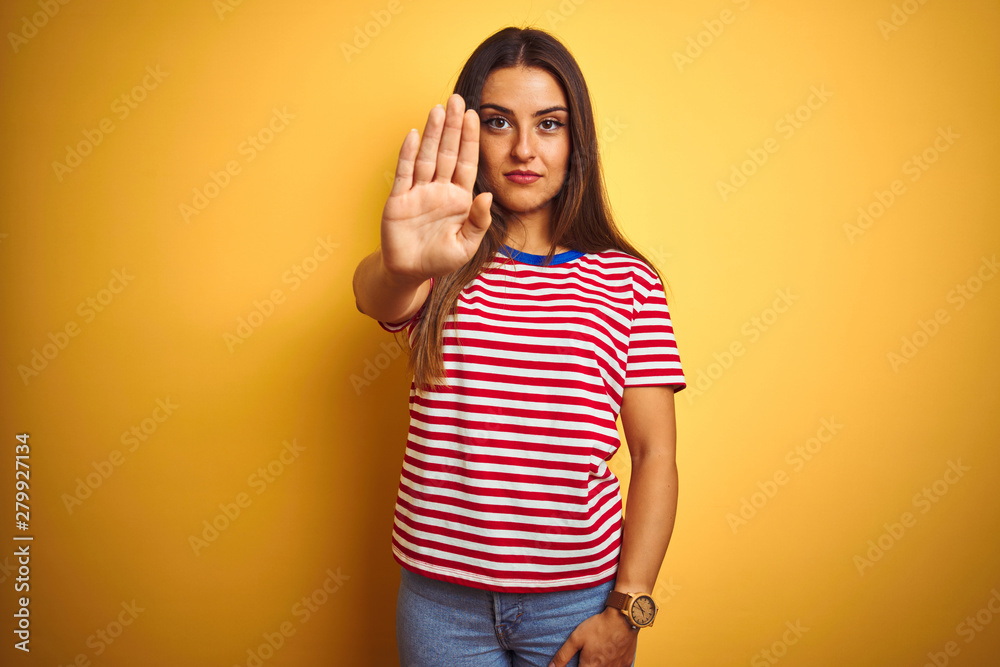 Young beautiful woman wearing striped t-shirt standing over isolated yellow background doing stop sing with palm of the hand. Warning expression with negative and serious gesture on the face. Young beautiful woman wearing striped t-shirt standing over isolated yellow background doing stop sing with palm of the hand. Warning expression with negative and serious gesture on the face.