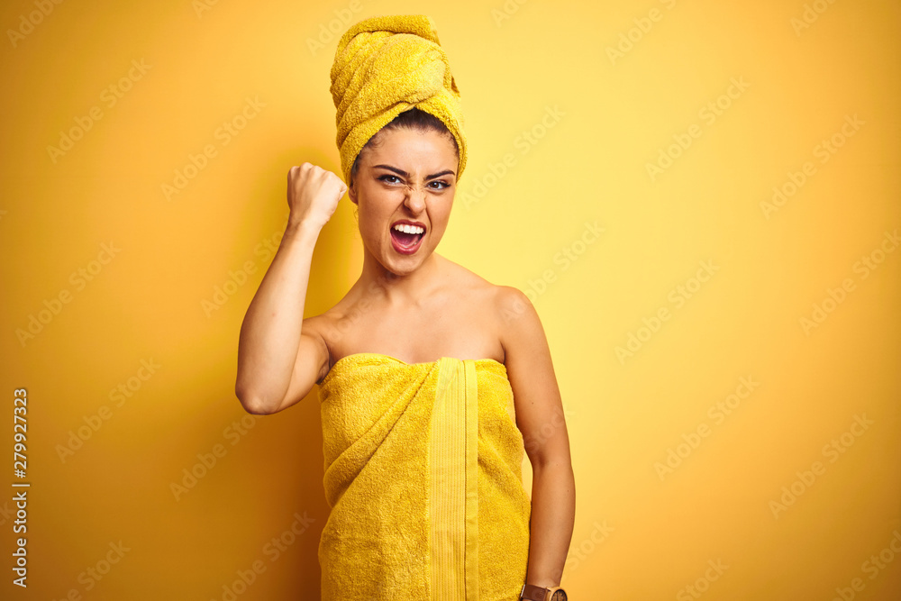 Young beautiful woman wearing towel after shower over isolated yellow background angry and mad raising fist frustrated and furious while shouting with anger. Rage and aggressive concept.