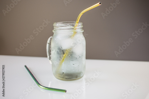 mason jar with ice cold water and reusable metal steel golden eco friendly straw on white table with another green bright straw on a side.