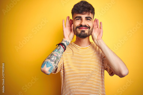 Young man with tattoo wearing striped t-shirt standing over isolated yellow background Trying to hear both hands on ear gesture, curious for gossip. Hearing problem, deaf