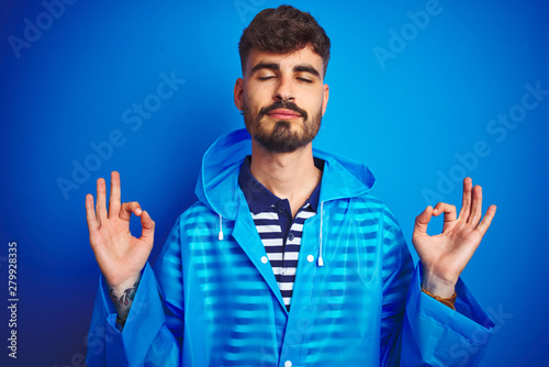 Young handsome man wearing rain coat standing over isolated blue background relax and smiling with eyes closed doing meditation gesture with fingers. Yoga concept.